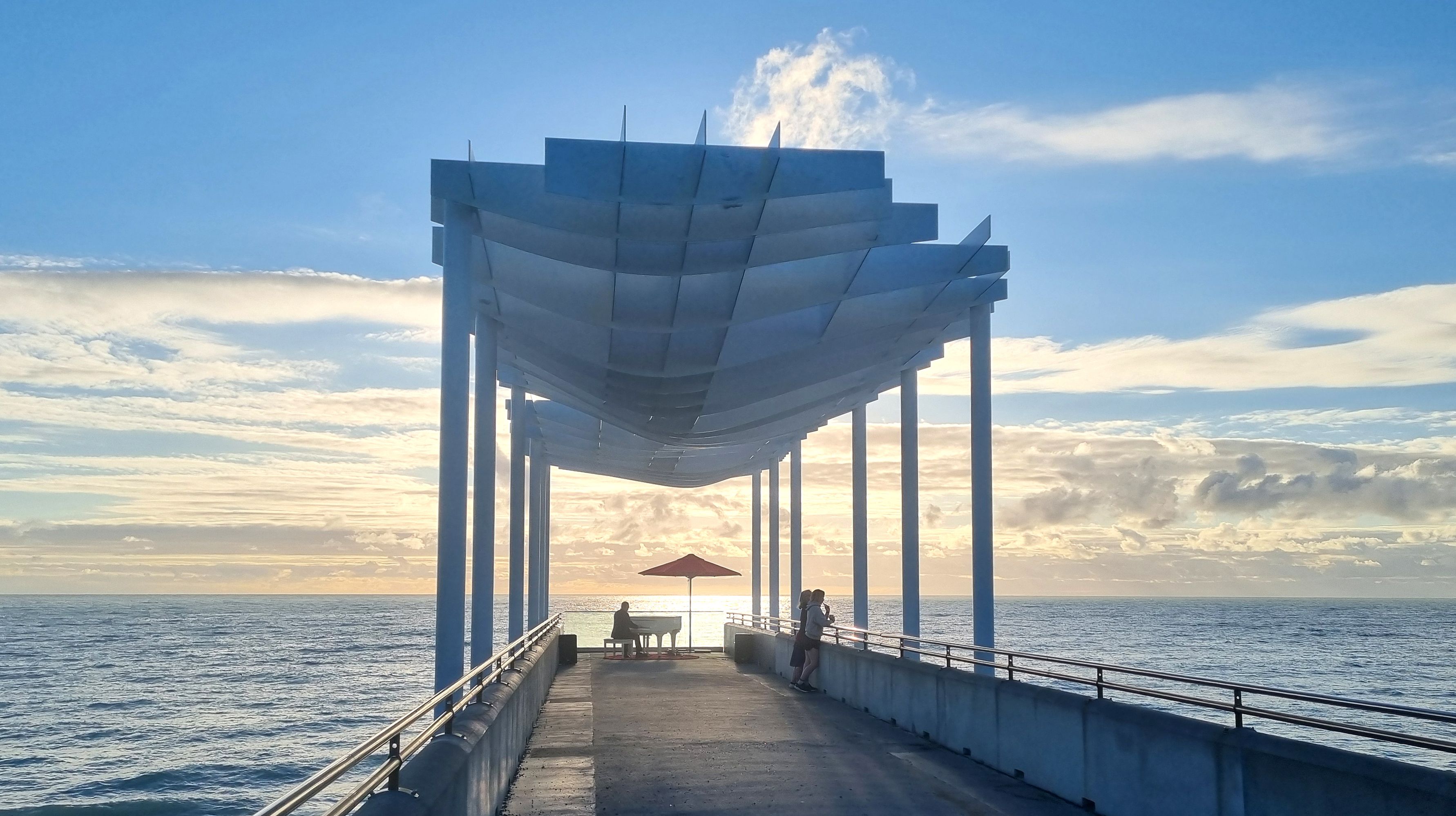 A distant shot looking down a pier at the piano silhouetted against the setting sun
